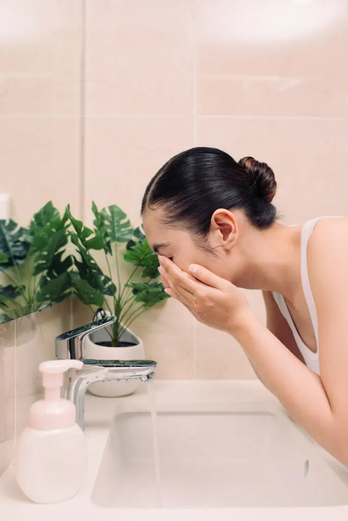 Woman washing her face with water above bathroom sink.