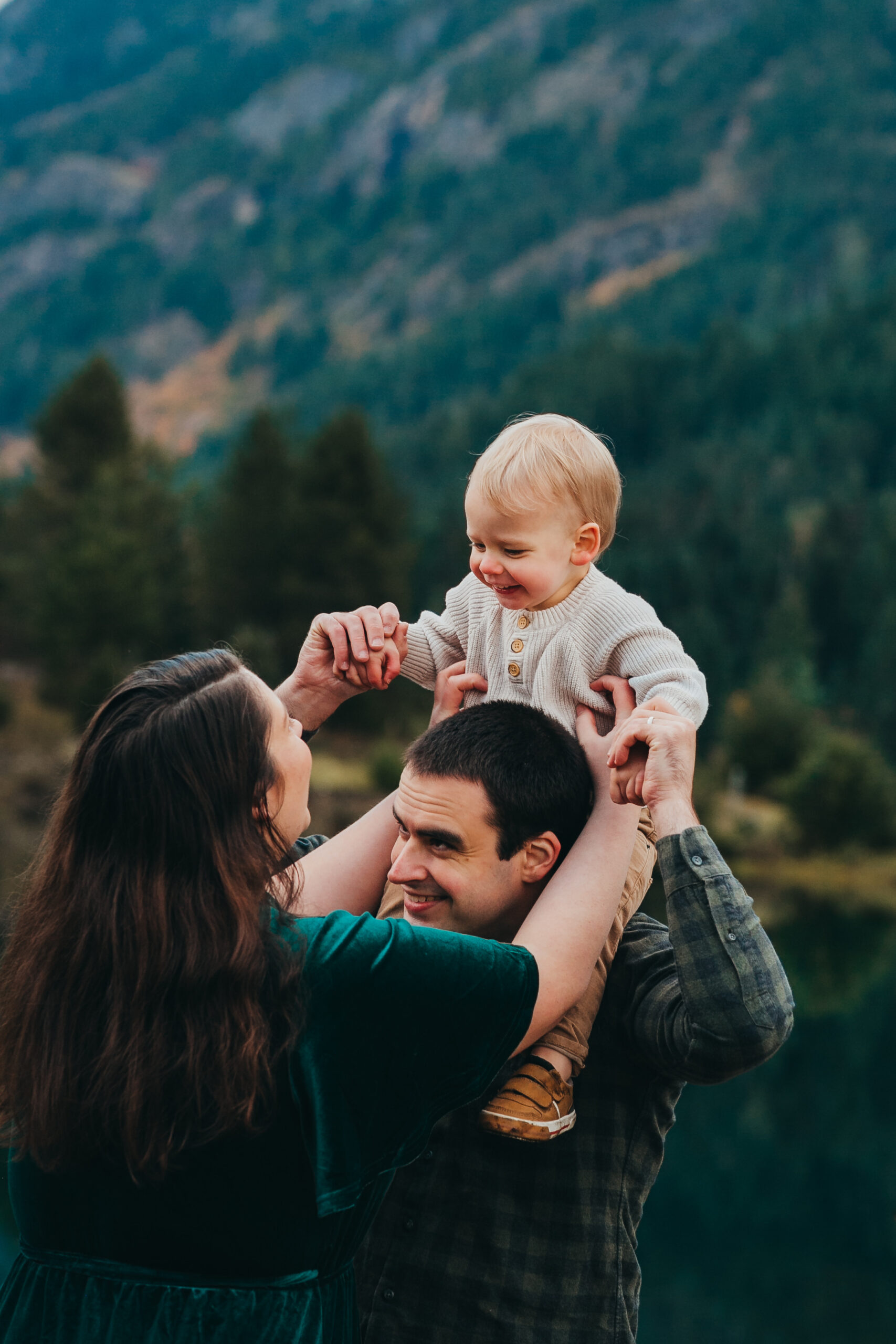 toddler on dad's shoulders while mom tickles - mountain backdrop