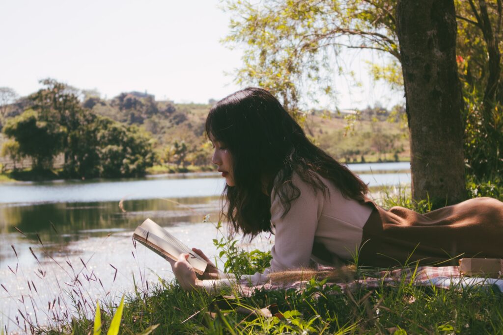 7 Striking Comparisons of Life in Your 20s vs 30sA woman lays on her stomach on a blanket, reading a book in the shade of trees along a river edge.