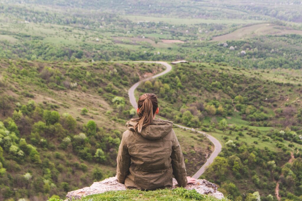 7 Striking Comparisons of Life in Your 20s vs 30sA woman sits on a mountain ledge looking over a green valley where a road winds away from her.